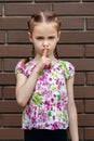 A serious child girl stands against the background of a brick wall and shows a gesture of silence with her hands Royalty Free Stock Photo