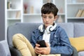 Serious child of a teenage boy in headphones sitting at home on the sofa, using the phone. Listens to music, watches Royalty Free Stock Photo