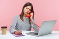 Serious concentrated brunette woman secretary answering call on landline telephone and writing down message, working on laptop