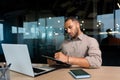 Serious concentrated businessman writing document while sitting in office using laptop for online learning, hispanic man Royalty Free Stock Photo