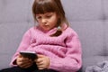 Serious concentrated dark haired female child wearing warm pink sweater, playing video game or checking social networks, sitting