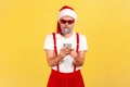 Serious concentrated gray bearded man in sunglasses and red santa claus hat holding smartphone and typing message or reading email