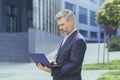 serious and concentrated gray haired boss working on laptop while standing outside modern office building, mature boss