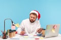 Serious concentrated man office worker sitting at workplace in santa claus hat, making notes at notepad holding smartphone in hand Royalty Free Stock Photo