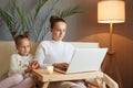 Serious concentrated mother looking at laptop? on sofa working online daughter sitting with mom and using smart phone waiting Royalty Free Stock Photo