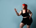 Serious concentrated young plus-size lady does exercises looking up with legs flexed and arms bent in elbows on blue background.