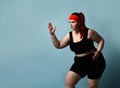 Serious concentrated young plus-size lady does exercises looking up with legs flexed and arms bent in elbows on blue background.