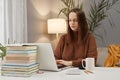 Serious concentrated young woman wearing brown sweater using laptop while working at home typing on keyboard writing article Royalty Free Stock Photo