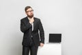 Serious confident thoughtful bearded young businessman in black suit are standing near his working place and holding his beard Royalty Free Stock Photo