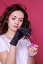 A serious curly brunette stands and holds in her hands a nail polish. A girl in a black glove holds a pink nail polish. Photoshoot