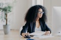 Serious curly businesswoman focused at display of computer, works on making project, surrounded with textbook and papers, wears