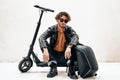 Serious and curly man wearing sunglasses posing in the bright studio while sitting on the scooter with a backpack Royalty Free Stock Photo