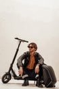 Serious and curly man wearing sunglasses posing in the bright studio while sitting on the scooter with a backpack Royalty Free Stock Photo