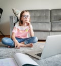 Serious cute little girl 8 years old in a striped T-shirt and jeans with glasses sits at home on a carpet in front of a laptop, Royalty Free Stock Photo