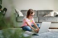 Serious cute little girl 8 years old in a striped T-shirt and jeans with glasses sits at home on a carpet in front of a laptop,