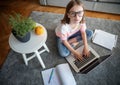 Serious cute little girl 8 years old in a striped T-shirt and jeans with glasses sits at home on a carpet in front of a laptop,