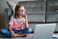 Serious cute little girl 8 years old in a striped T-shirt and jeans with glasses sits at home on a carpet in front of a laptop,