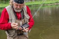 Serious elderly bearded fisherman on river bank holding rod and tackle box thinking what kind of fly fishing lure would be better