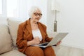 a serious, experienced, elderly woman is sitting working from home on the couch with a laptop on her lap and menacingly