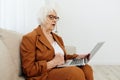 a serious, experienced, elderly woman is sitting working from home on the couch with a laptop on her lap and menacingly