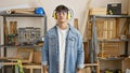 Serious-faced young hispanic man, a skilled carpenter, wearing glasses and headphones, standing relaxed in his woodworking