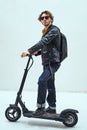 Serious and fashionable man wearing sunglasses posing in the bright studio while riding on the scooter with a backpack Royalty Free Stock Photo