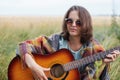 Serious female with attractive appearance wearing sunglasses sitting at meadow with acoustic guitar trying to play song. Young fem Royalty Free Stock Photo