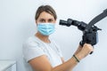 Serious female dentist in medical mask holding handles of dental microscope and looking aside working with device. Royalty Free Stock Photo