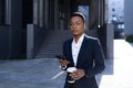 Serious female student thinking, holding and using phone, african american business woman with cup of hot drink walking on break Royalty Free Stock Photo