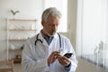Serious focused mature doctor using phone, looking at screen