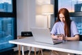 Serious focused young woman using phone and sitting at desk with laptop computer in apartment near window evening at Royalty Free Stock Photo