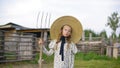 Serious girl in dress and wicker hat posing with hayfork in village