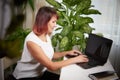 Serious girl with laptop and a notebook is working at a table in cozy room with flowers. A middle-aged business woman