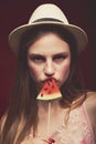 Serious girl with pink make up, wearing jeans, hat and top, posing at red studio background, holding slice watermelon on stick and Royalty Free Stock Photo