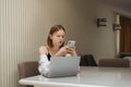 Serious girl sitting with laptop at desk in apartment and embarrassed looking in smartphone screen.Beautiful business woman in Royalty Free Stock Photo