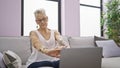 Serious grey-haired senior woman, focused on using laptop, sitting on sofa stretching tired arms at her cozy home living room Royalty Free Stock Photo