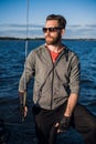 Serious handsome young male yacht captain in sunglasses wearing gloves staying on boat deck and looking into distance