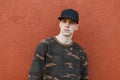 Serious handsome young man in fashionable green camouflage clothing in a black stylish cap stands near a vintage red wall outdoors Royalty Free Stock Photo