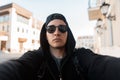 Serious handsome young man hipster in trendy dark sunglasses with a stylish cap in a black hoodie makes a selfie in the city Royalty Free Stock Photo