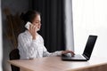 Serious hispanic businesswoman using laptop computer talking on mobile phone consult client sit at desk, focused female Royalty Free Stock Photo