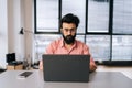 Serious Indian business man in casual clothes sitting at desk with laptop looking at camera in light coworking office by Royalty Free Stock Photo