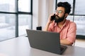 Serious Indian business man in glasses working at laptop computer and talking on mobile phone sitting at desk by window. Royalty Free Stock Photo