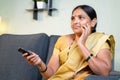 Serious Indian woman in saree watching TV serials at home while sitting on sofa by holding remote during leisure time