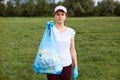 Serious lady in baseball cap holding garbage bag full of trash in hands, showing it to people, calls on to clean up our planet, Royalty Free Stock Photo