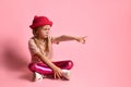 Little blonde girl in hat, t-shirt, leggings and sneakers. She looking frowned, sitting on floor with crossed legs against pink Royalty Free Stock Photo