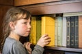 Serious little girl is taking a yellow book from the bookshelf. The child gets a textbook from the shelf of the old library. Royalty Free Stock Photo