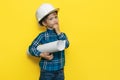 Serious little kid boy engineer or architect in a protective helmet holding construction plan over yellow background. Royalty Free Stock Photo