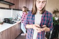 Serious long-haired young man standing in the kitchen uses his mobile phone to write SMS or order a service from the Royalty Free Stock Photo