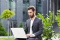 Serious male businessman working on laptop at lunchtime sitting on bench near office in business suit Royalty Free Stock Photo