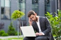 Serious male businessman working on laptop at lunchtime sitting on bench near office in business suit Royalty Free Stock Photo